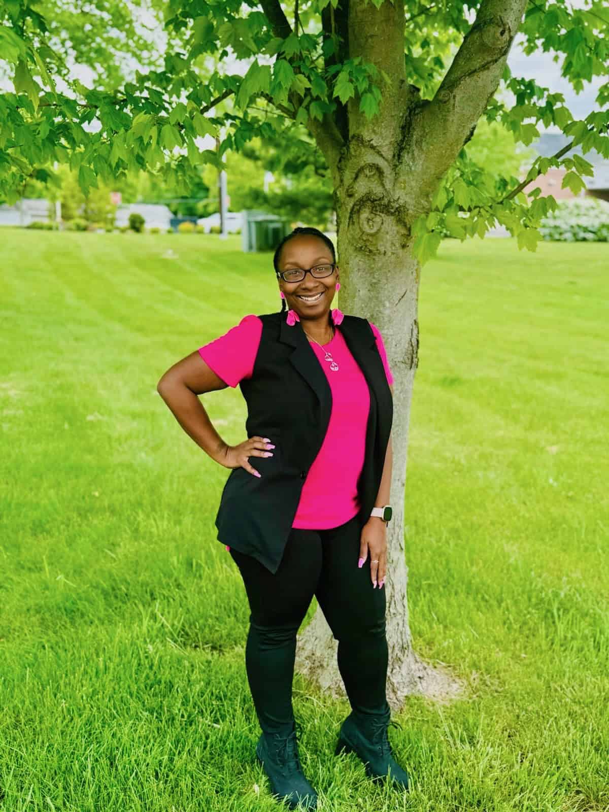 Woman in black and pink outfit standing confidently by a tree on a grassy lawn, smiling at the camera.